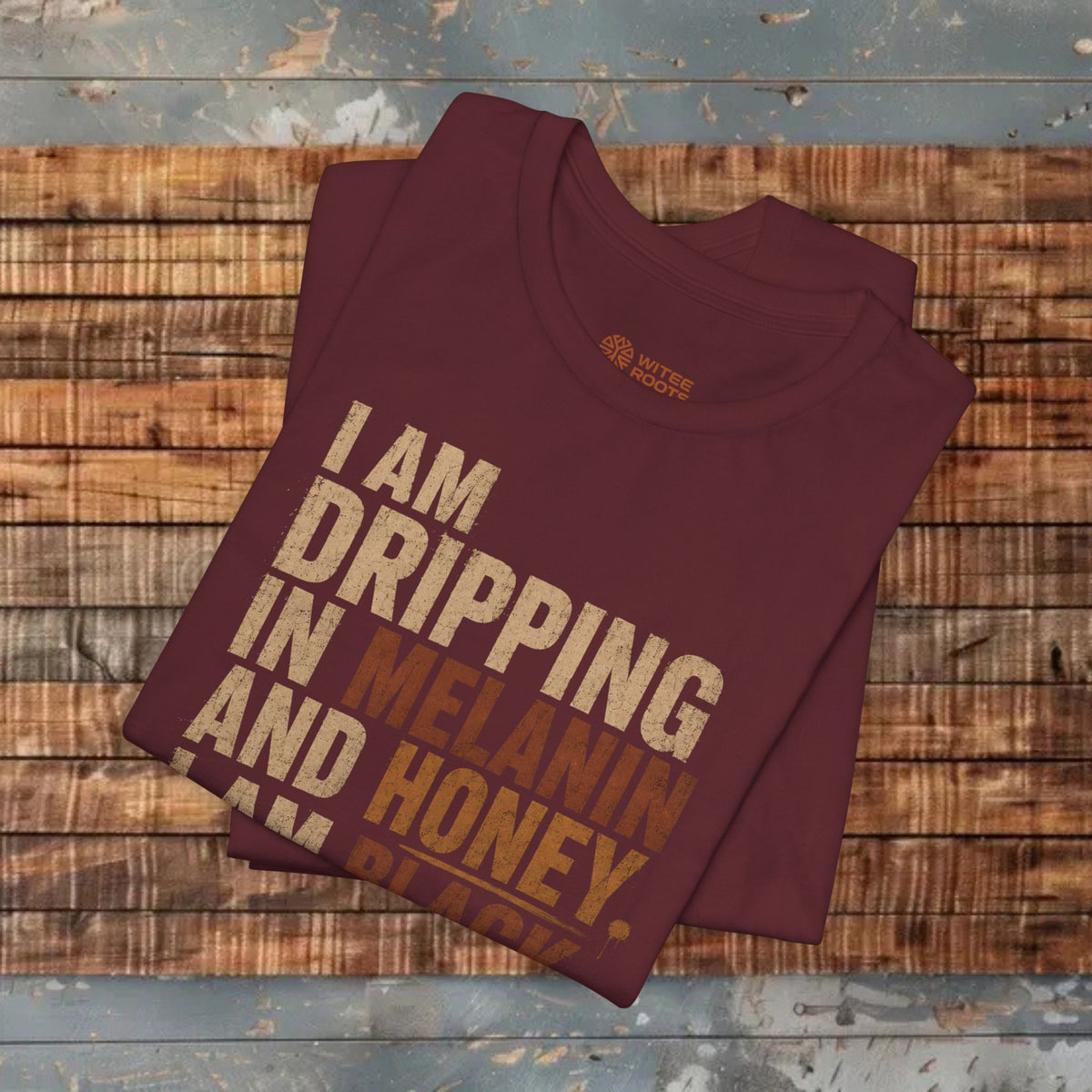 A folded maroon t-shirt with a humorous text graphic reading 'I AM DRIPPING IN MELANIN AND HONEY. I AM BLACK.