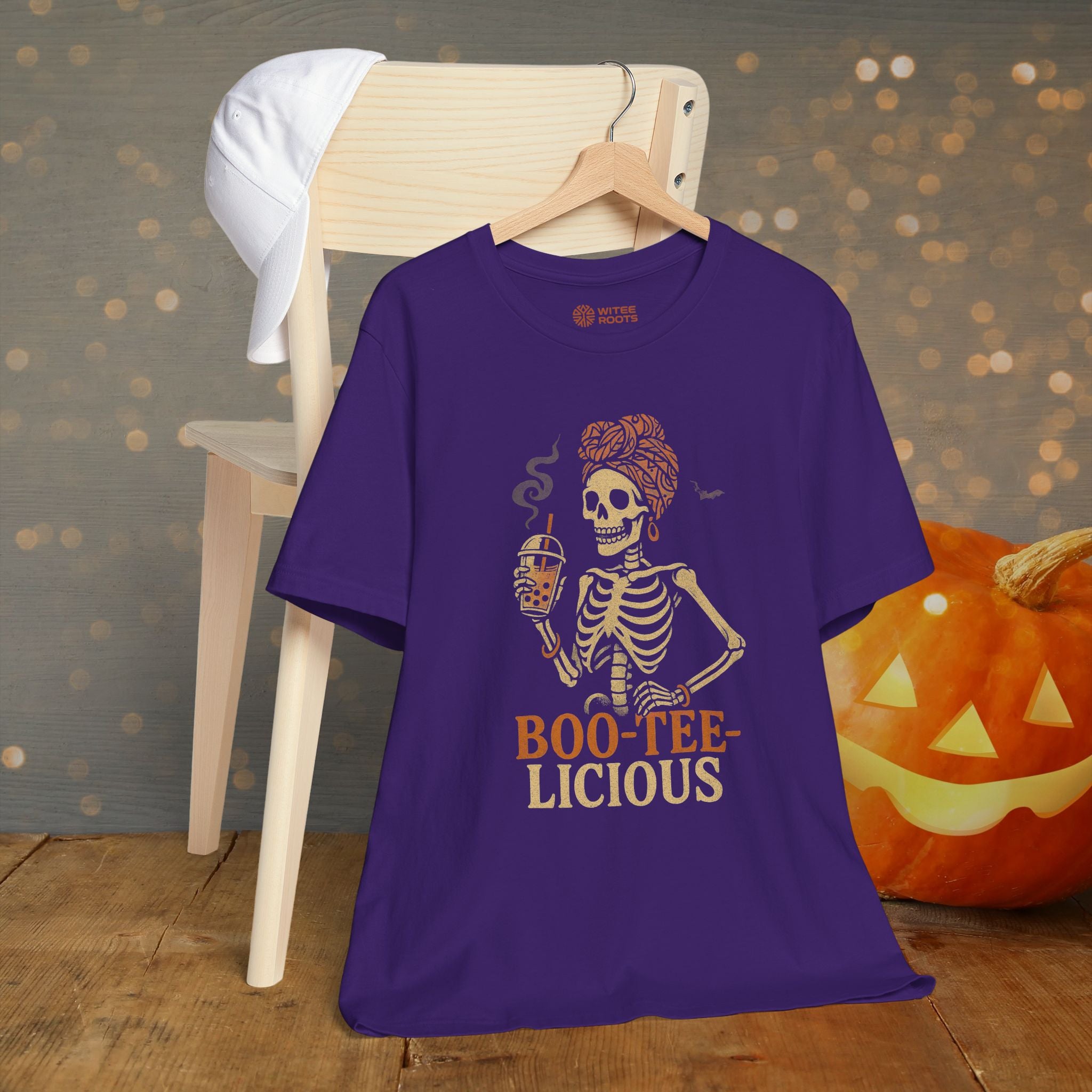 Purple t-shirt with skeleton graphic and 'BOO-TEE-LICIOUS' text, displayed on a hanger with a pumpkin in the background.