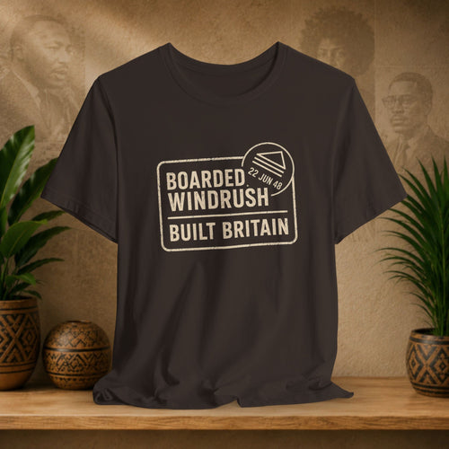 Brown t-shirt with 'Boarded Windrush Built Britain' text on a wooden surface with plants and a vintage-style portrait in the background.