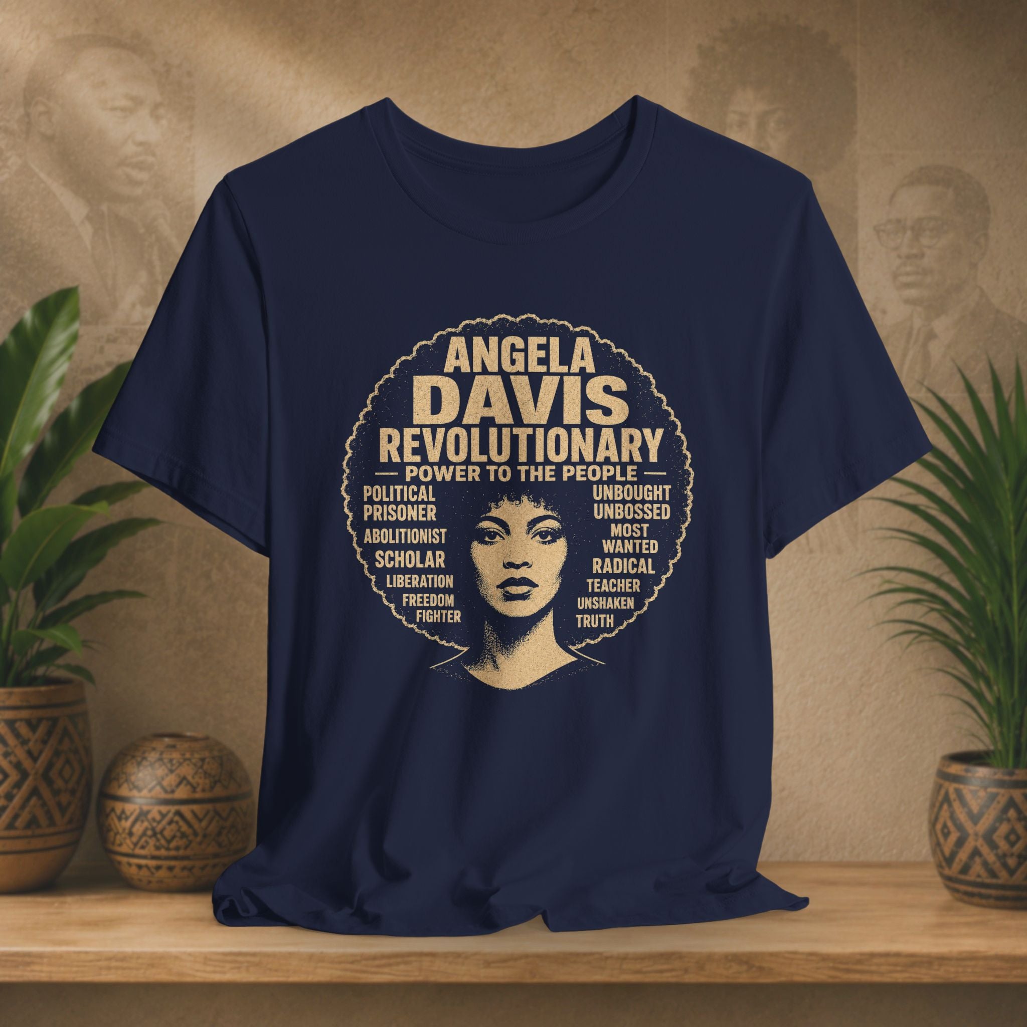 A navy blue graphic t-shirt featuring a circular design with the text 'ANGELA DAVIS REVOLUTIONARY' and a portrait of Angela Davis, displayed on a wooden shelf with potted plants in the background.