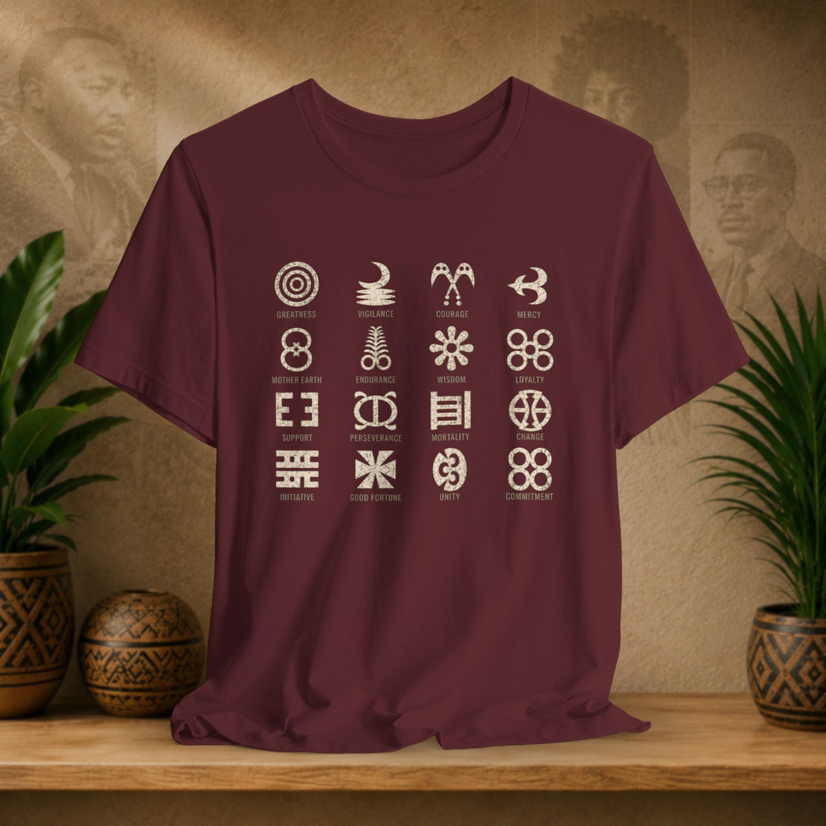 A maroon short-sleeve t-shirt with a grid of 18 white Aboriginal symbols and their names printed on the front, displayed on a wooden shelf with potted plants and a textured background.