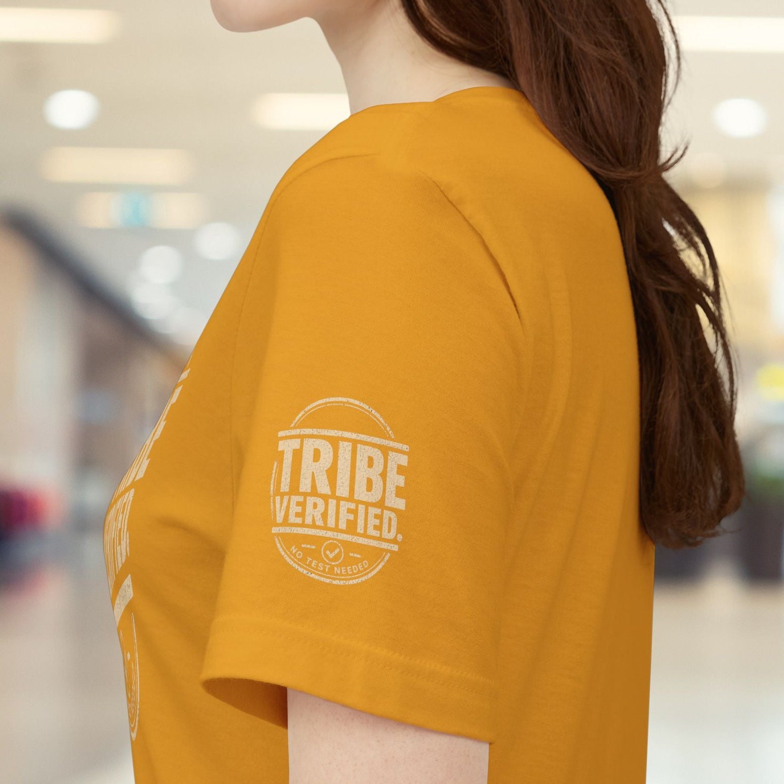 Person wearing a mustard yellow t-shirt with 'TRIBE VERIFIED' logo in a blurred indoor setting
