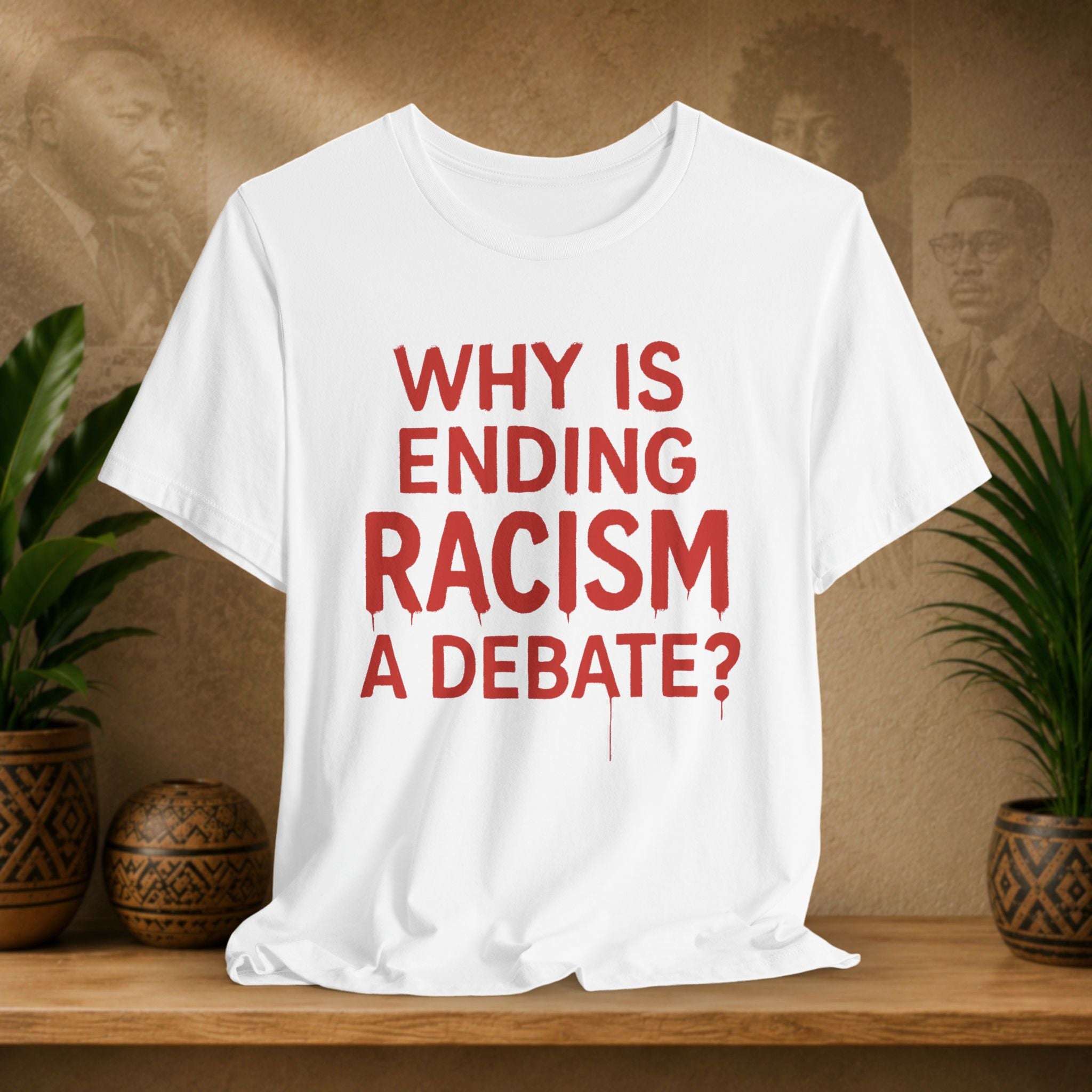 White t-shirt with red text 'WHY IS ENDING RACISM A DEBATE?' on a wooden surface with plants and a textured wall in the background.