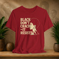 Red t-shirt with 'Black Don't Crack, It Resists' text and graphic on a wooden surface with plants and decorative items.