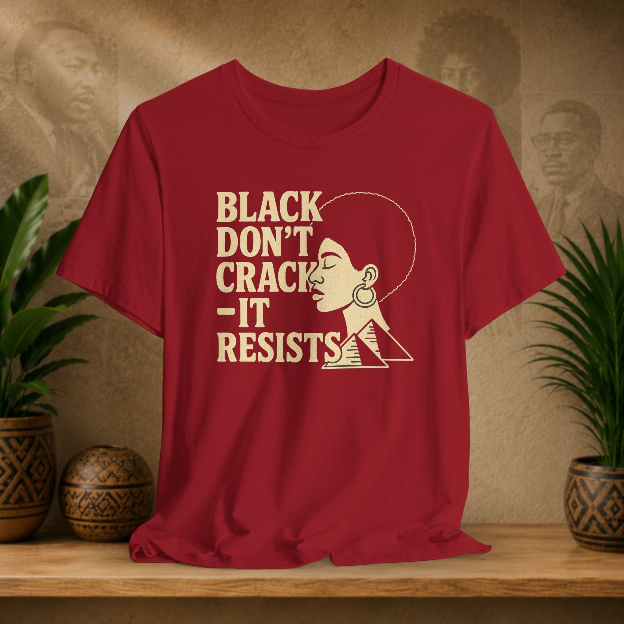 Red t-shirt with 'Black Don't Crack, It Resists' text and graphic on a wooden surface with plants and decorative items.