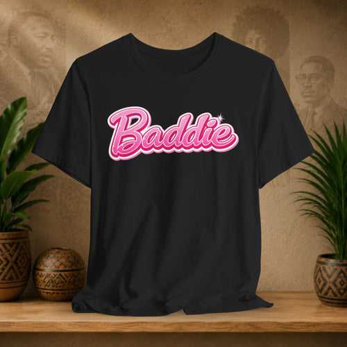 A black short-sleeve t-shirt with 'Baddie' written in pink cursive script across the chest, styled on a wooden shelf with potted plants and a textured background.