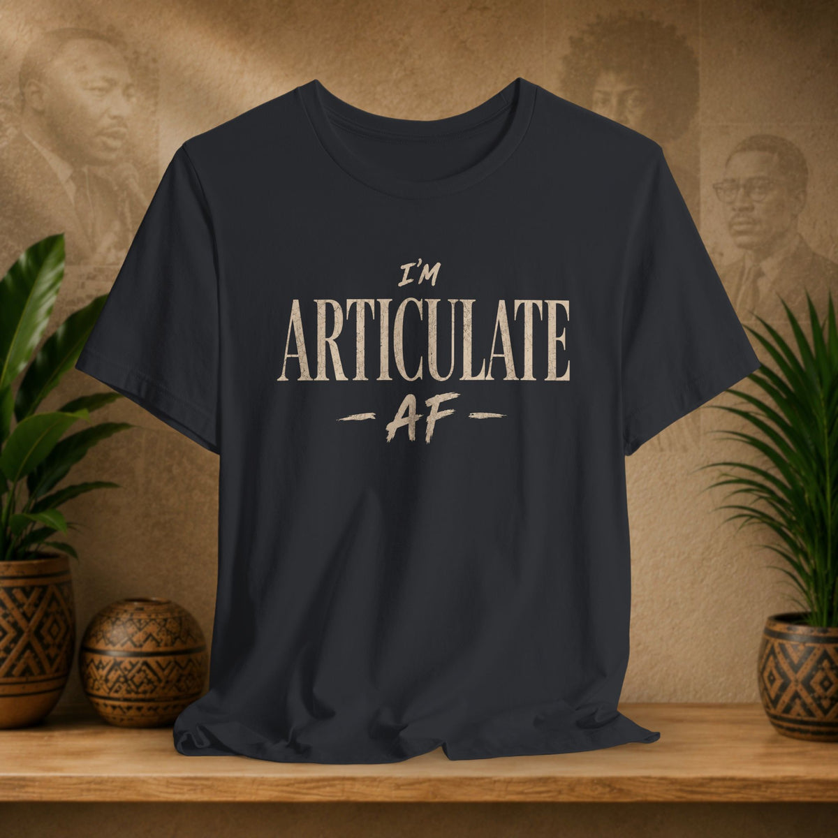 A black crewneck t-shirt with the text 'I'M ARTICULATE AF' printed in a distressed beige font, displayed on a wooden shelf with potted plants and a textured background.