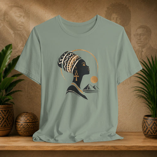 Sage green t-shirt with a graphic of a woman's profile, headwrap, and mountains on a wooden surface with plants and decorative items.