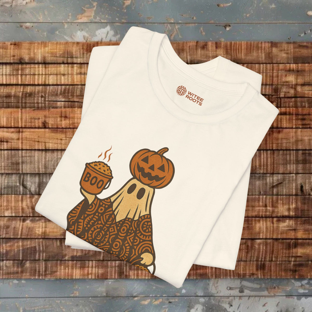 A folded white t-shirt with a Halloween graphic of a ghost in a paisley pattern holding a pumpkin cup, displayed on a rustic wooden background. The inner collar shows the 'Rustic Routes' brand logo.