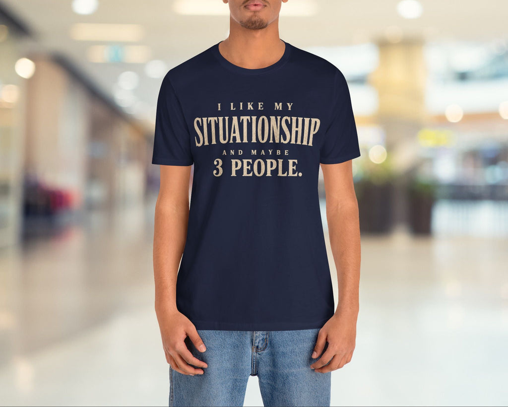 A man wearing a navy blue t-shirt with a beige graphic print that reads 'I LIKE MY SITUATIONSHIP AND WASTE 3 PEOPLE.' standing in a blurred indoor hallway.