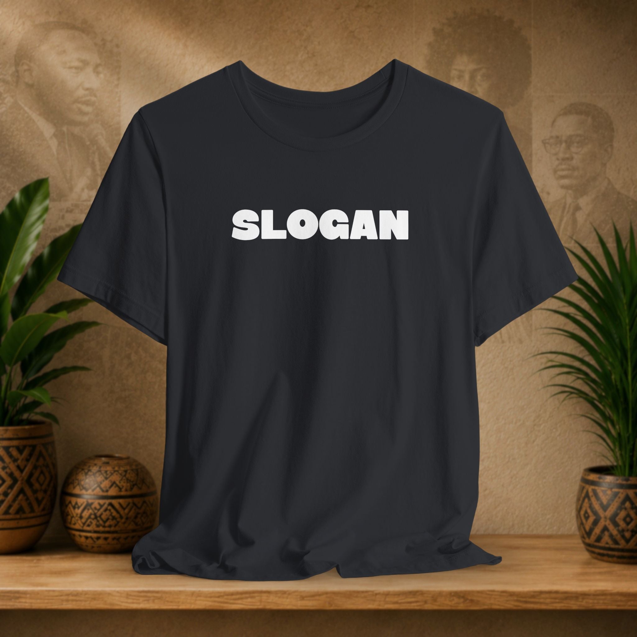 A dark charcoal gray t-shirt with the word 'SLOGAN' printed in bold white capital letters across the chest, displayed on a wooden shelf with decorative plants and textured background.