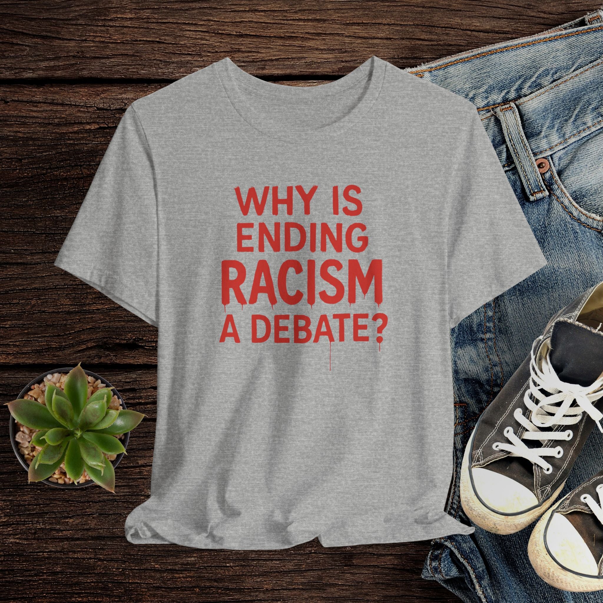 Gray t-shirt with red text 'WHY IS ENDING RACISM A DEBATE?' on a wooden surface with jeans and sneakers.