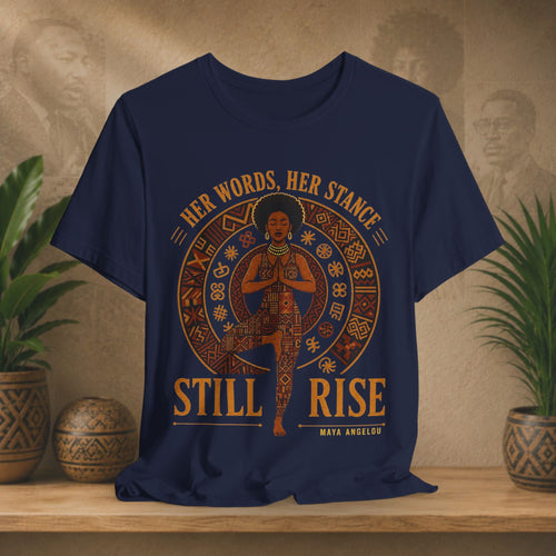 A navy blue graphic t-shirt displayed on a wooden shelf with potted plants in the background. The shirt features a circular design with a central figure in a yoga pose, surrounded by the text 'HER WORDS, HER STANCE' at the top and 'STILL RISE' at the bottom, with 'MAYA ANGELUS' written underneath.