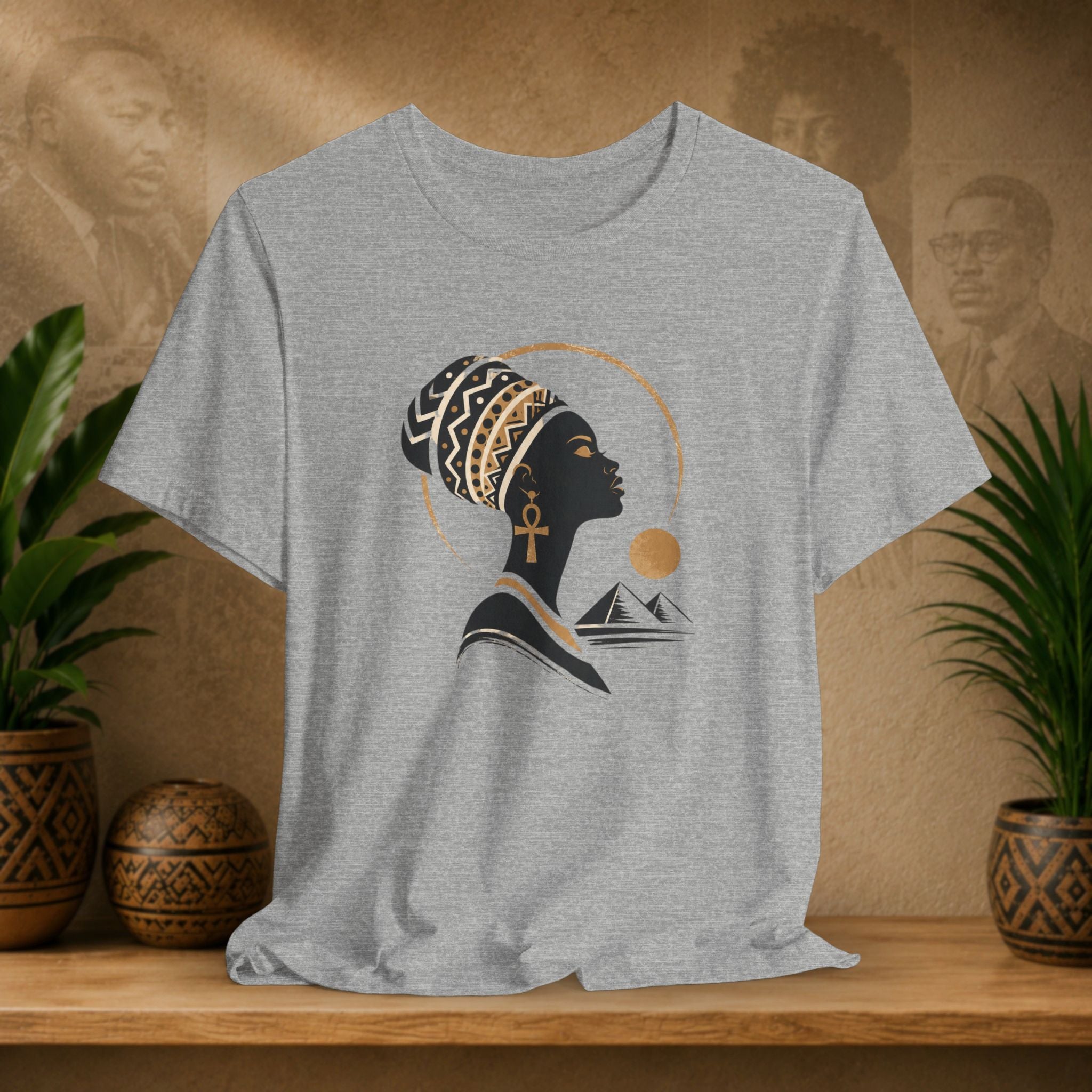 Gray t-shirt with a graphic of a woman's profile, an ankh, and mountains on a wooden surface with plants and a textured wall.