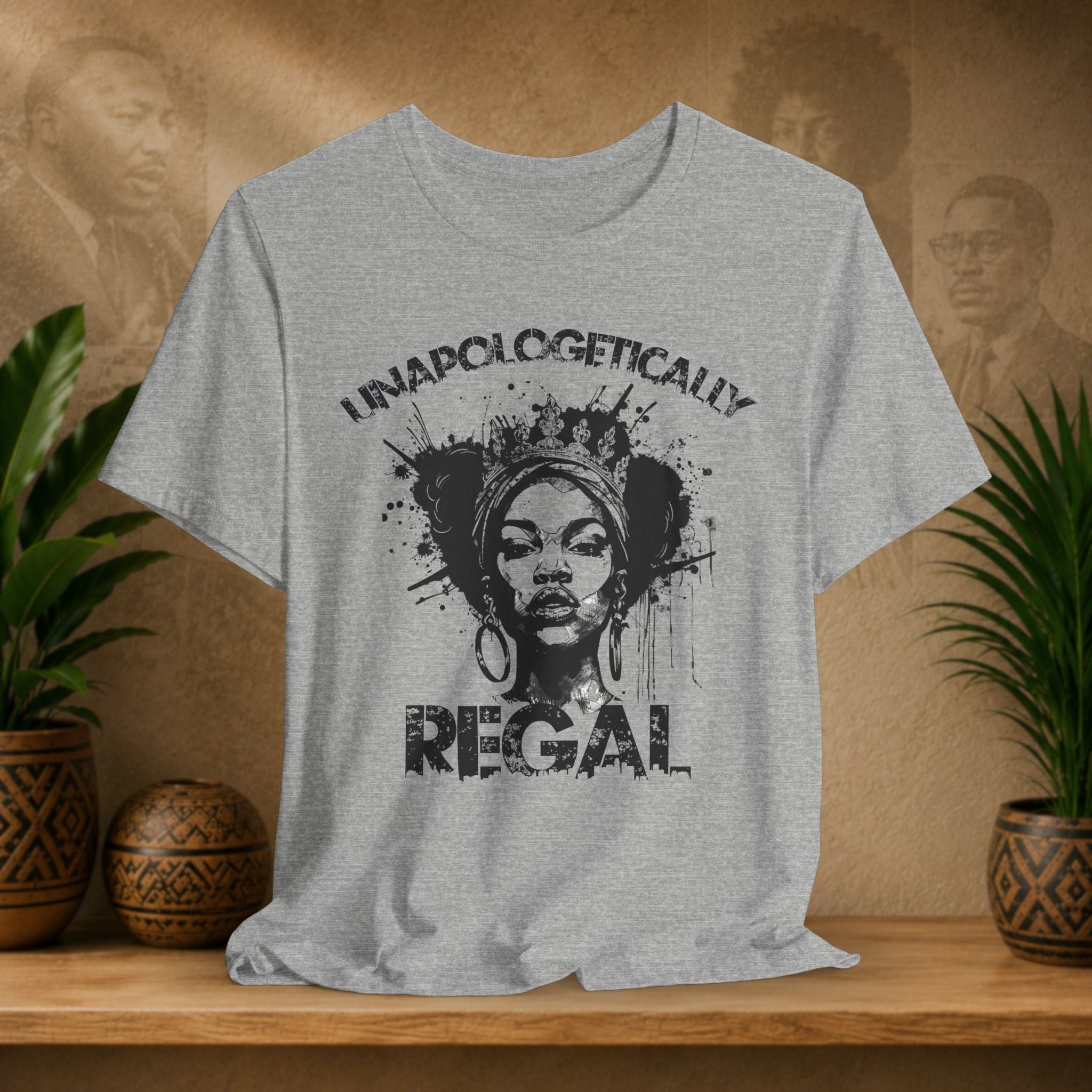 A light heather gray t-shirt with a black graphic print of a woman's face and the text 'UNAPOLOGETICALLY REGAL', displayed on a wooden shelf with potted plants in the background.