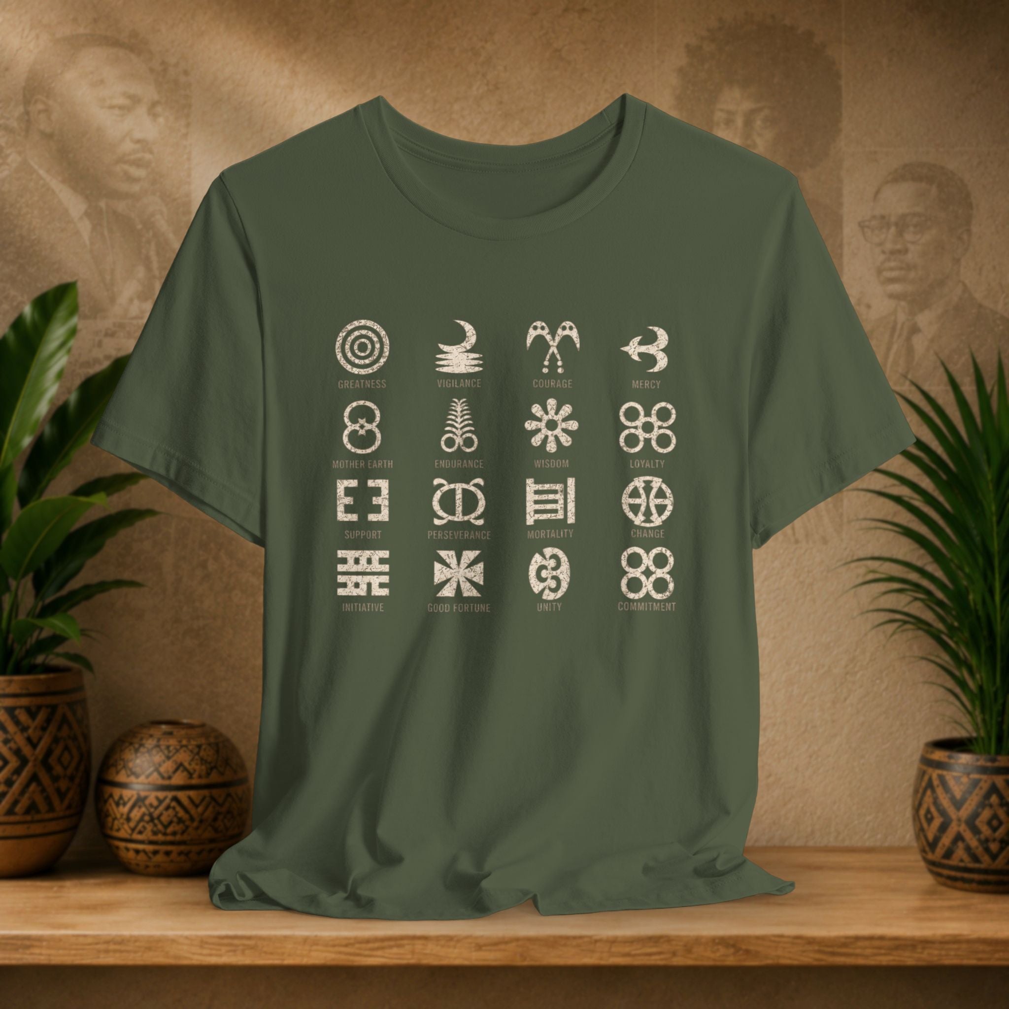 An olive green short-sleeve t-shirt with a 4x4 grid of white tribal and spiritual symbols on the front, displayed on a wooden shelf with potted plants and a textured background.