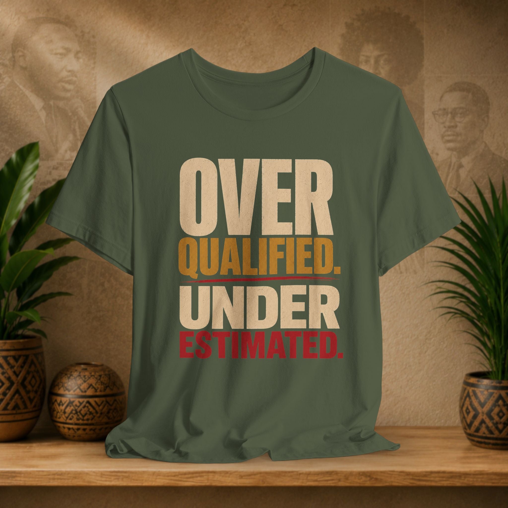 An olive green graphic t-shirt displayed on a wooden shelf, flanked by potted plants and decorative vases against a textured beige background with faint portraits. The shirt features the text 'OVER QUALIFIED.