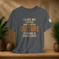 A dark gray graphic t-shirt displayed on a wooden shelf, flanked by potted green plants and decorative baskets. The shirt features the text 'I LIKE MY COFFEE HOW I LIKE MY CULTURE STRONG & UNFILTERED' in cream and orange lettering, with a small coffee cup graphic on the left sleeve.