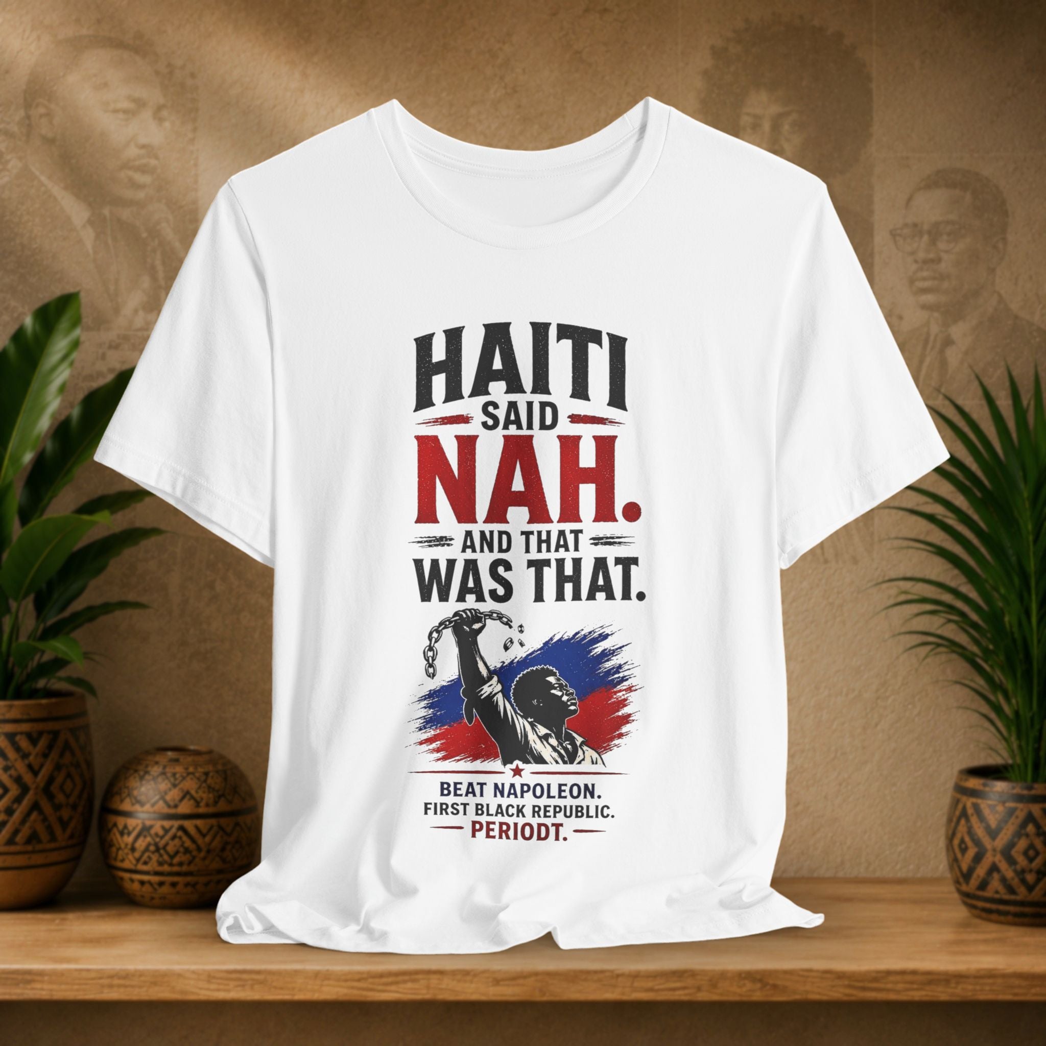 White t-shirt with Haitian text and graphics on a wooden shelf with plants and a textured wall.