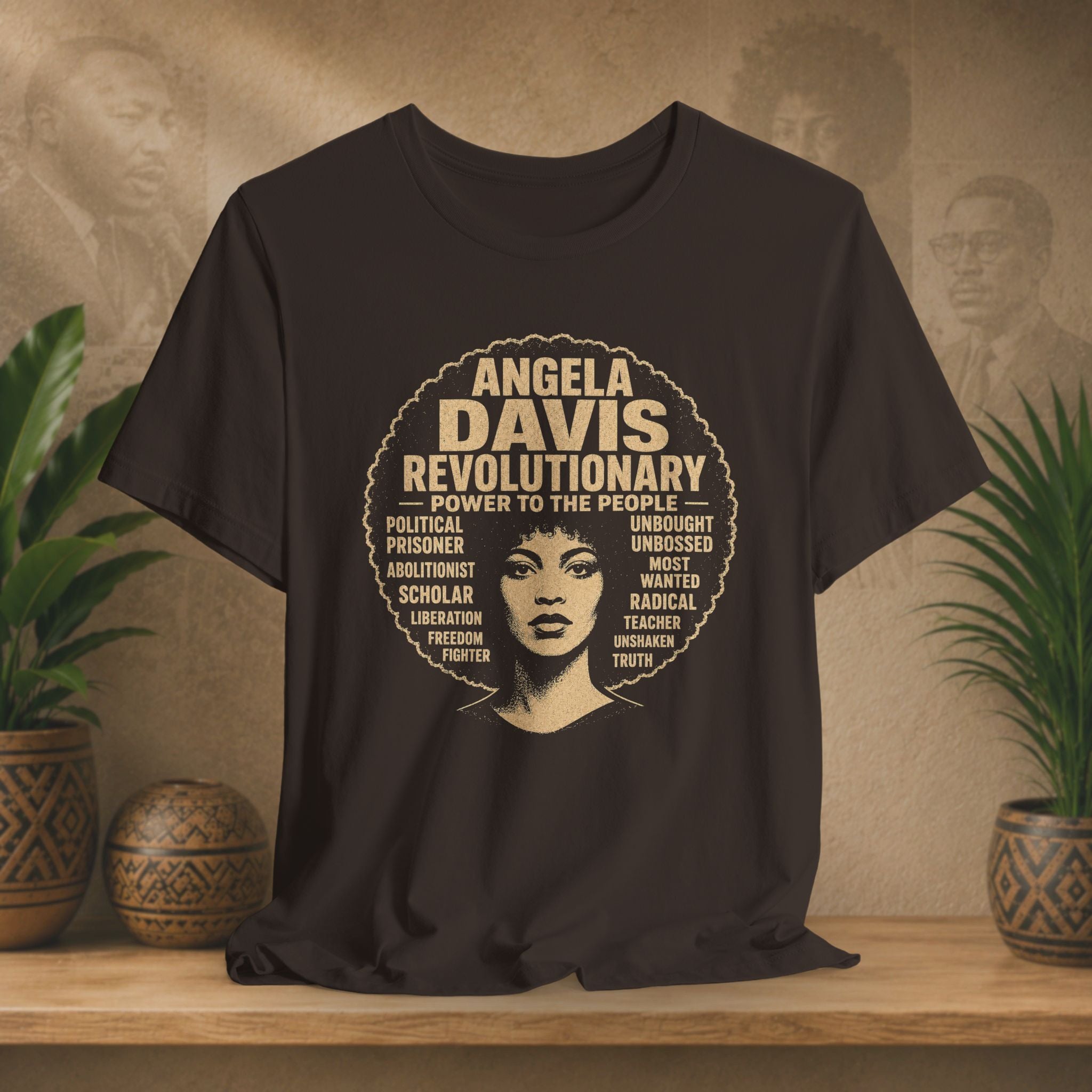 A brown short-sleeve graphic T-shirt displayed on a wooden surface against a warm beige wall with potted plants. The shirt features a large circular graphic with the text 'ANGELA DAVIS REVOLUTIONARY' and a stylized portrait of Angela Davis in the center, surrounded by smaller text.