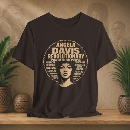 A brown short-sleeve graphic T-shirt displayed on a wooden surface against a warm beige wall with potted plants. The shirt features a large circular graphic with the text 'ANGELA DAVIS REVOLUTIONARY' and a stylized portrait of Angela Davis in the center, surrounded by smaller text.
