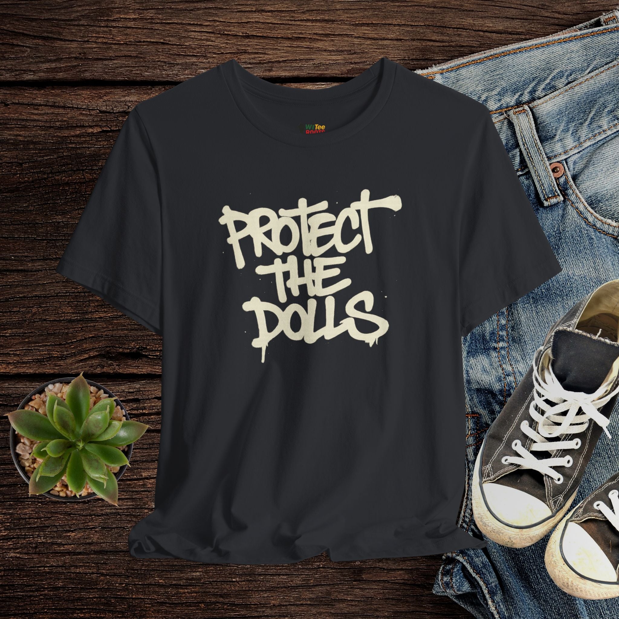 Black t-shirt with 'Protect the Dolls' text on a wooden surface with jeans and sneakers.