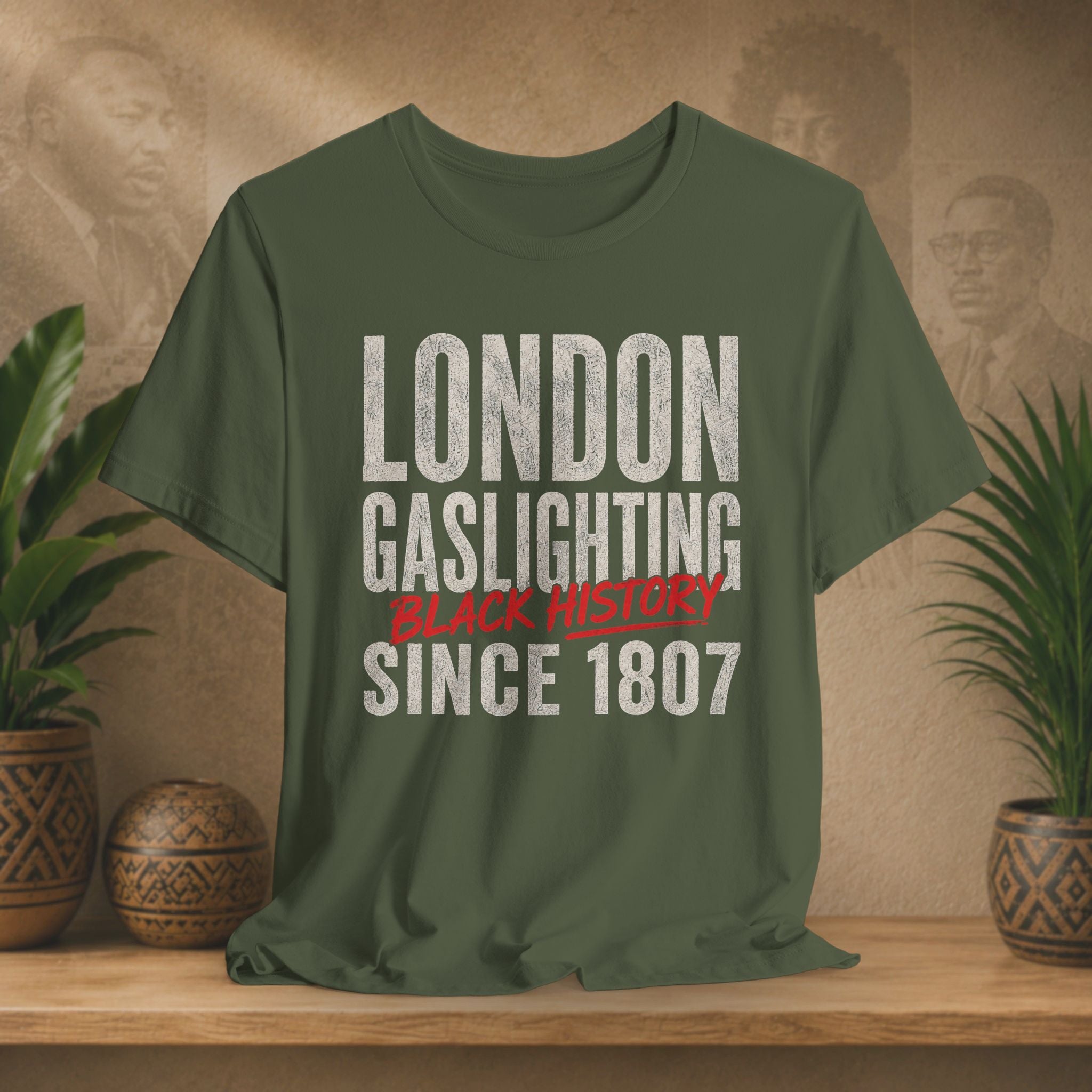 Green t-shirt with 'London Gaslighting Black History Since 1807' text on a wooden surface with plants and a wall in the background.