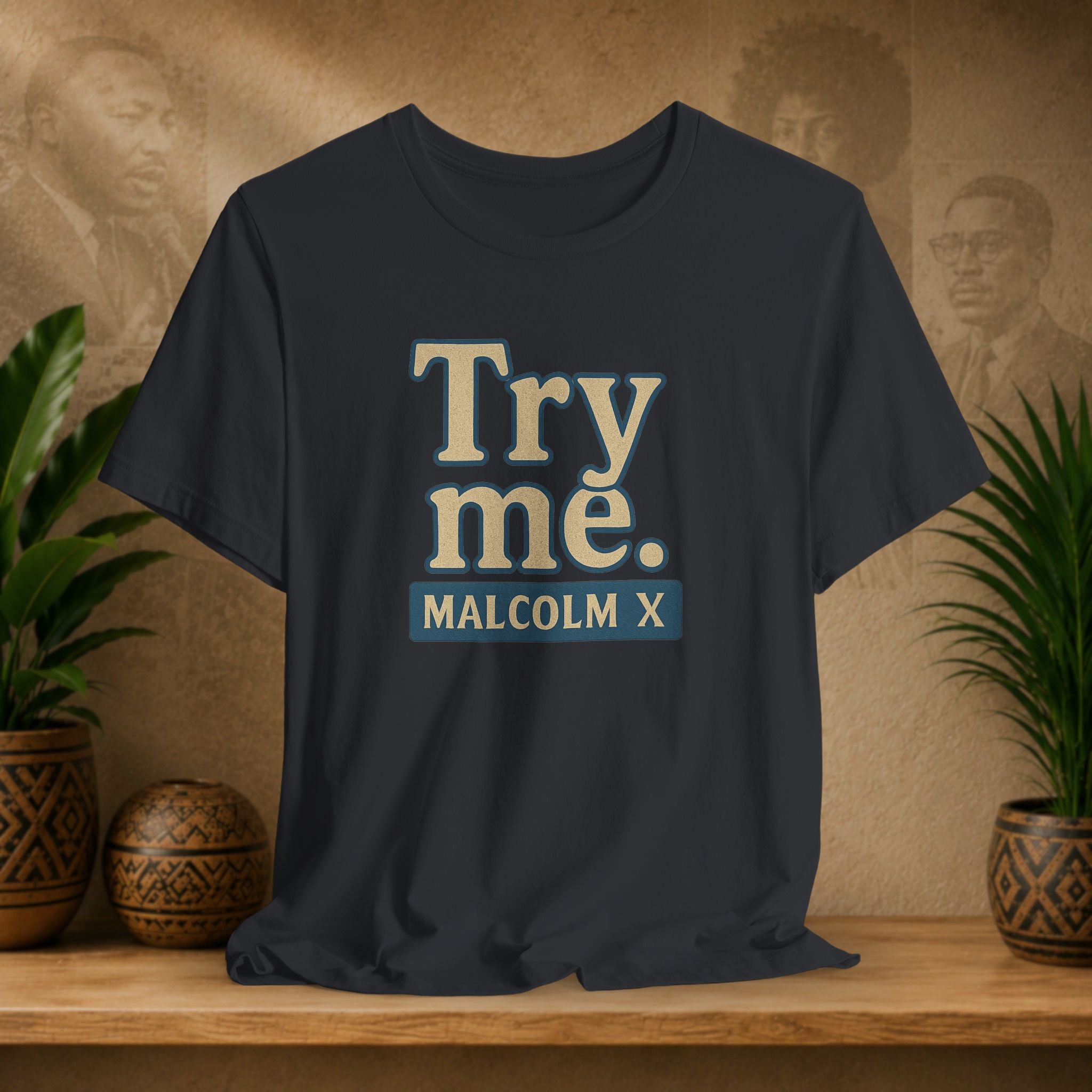 Black t-shirt with 'Try me. MALCOLM X' text on a wooden surface with plants and a blurred background.