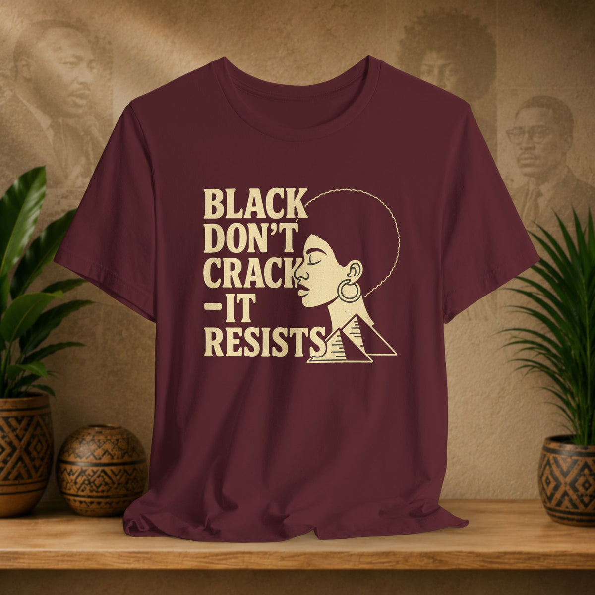 A maroon short-sleeve graphic T-shirt displayed on a wooden surface, featuring the text 'BLACK DON'T CRACK - IT RESISTS' in beige next to a stylized profile illustration of a woman with an afro. The background shows a warm textured wall with faded portraits and potted plants on both sides.