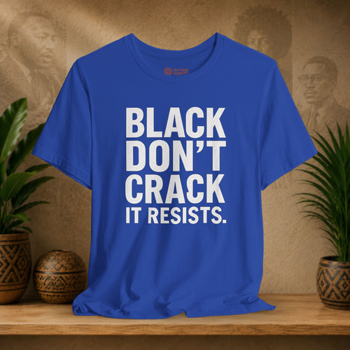 Blue t-shirt with 'BLACK DON'T CRACK IT RESISTS.' text on a wooden surface with plants and decorative items.