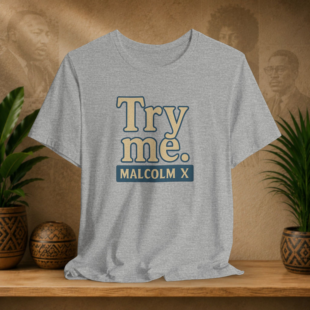 Gray t-shirt with 'Try me. MALCOLM X' text on a wooden shelf with plants and a blurred background.