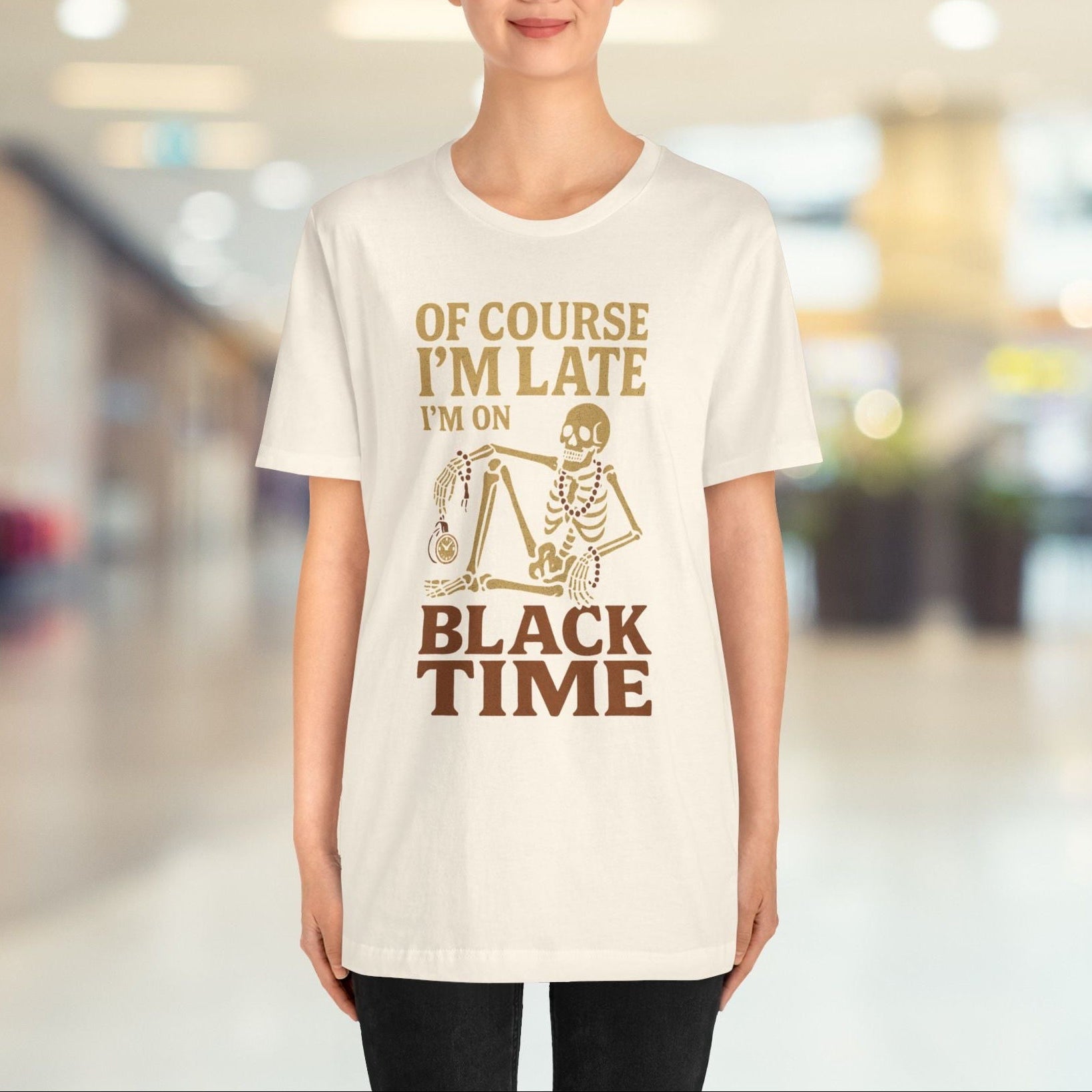 A woman stands in a brightly lit indoor hallway wearing a white graphic T-shirt with brown text reading 'OF COURSE I'M LATE I'M ON BLACK TIME' and a small skeleton illustration.