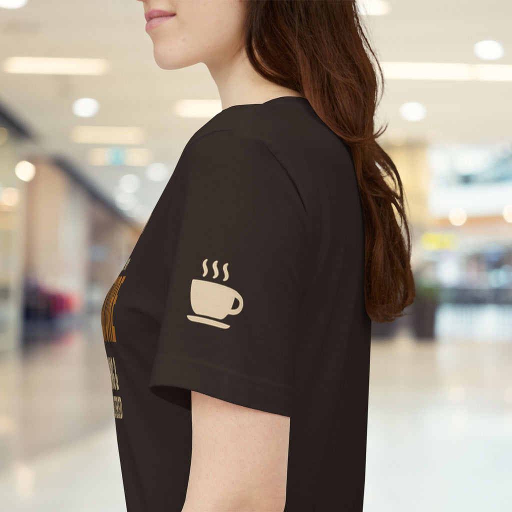 A woman wearing a brown short-sleeve t-shirt with a beige coffee cup graphic on the sleeve, photographed in a blurred indoor shopping mall setting.
