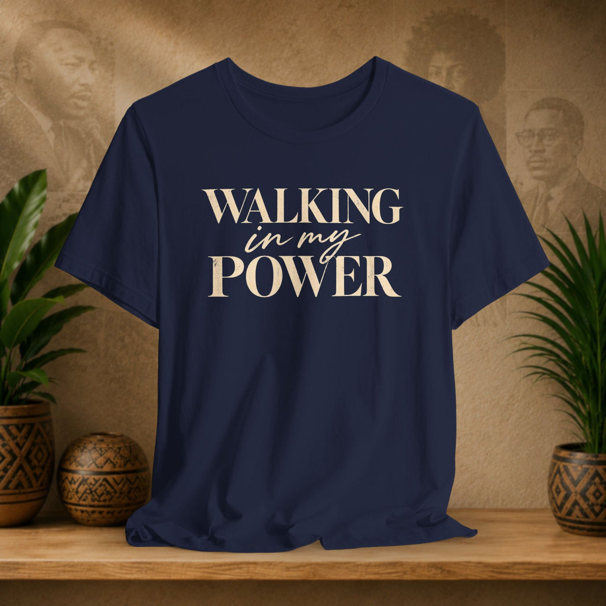A navy blue short-sleeve t-shirt with the text 'WALKING in my POWER' printed in beige on the front, displayed on a wooden shelf with potted plants and a textured background.
