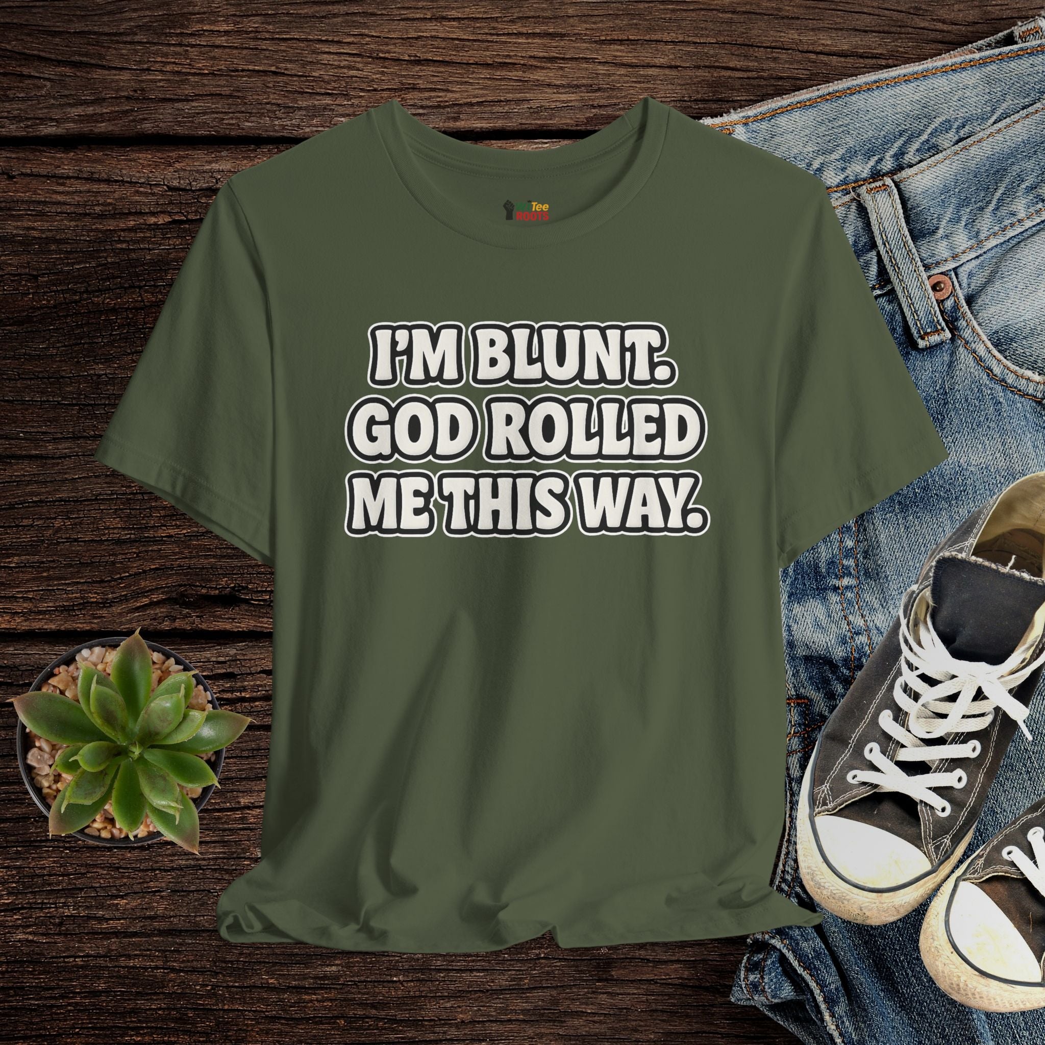 Green t-shirt with text 'I'M BLUNT. GOD ROLLED ME THIS WAY.' on a wooden surface with jeans and sneakers.