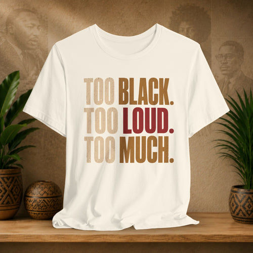 A beige short-sleeve graphic T-shirt displayed on a wooden surface, featuring the text 'TOO BLACK. TOO LOUD.