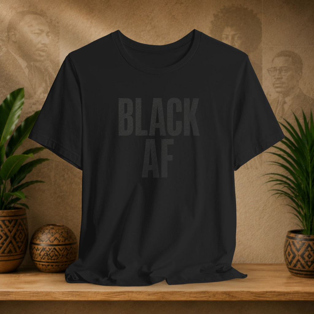A black short-sleeve crewneck t-shirt with the words 'BLACK AF' printed across the chest in a faded dark gray font. The shirt is displayed on a wooden shelf flanked by potted green plants and decorative ceramic vases against a textured beige wall with faint portraits.