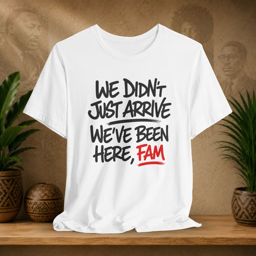 White t-shirt with text 'WE DIDN'T JUST ARRIVE WE'VE BEEN HERE, FAM' on a wooden surface with plants and a textured wall background.