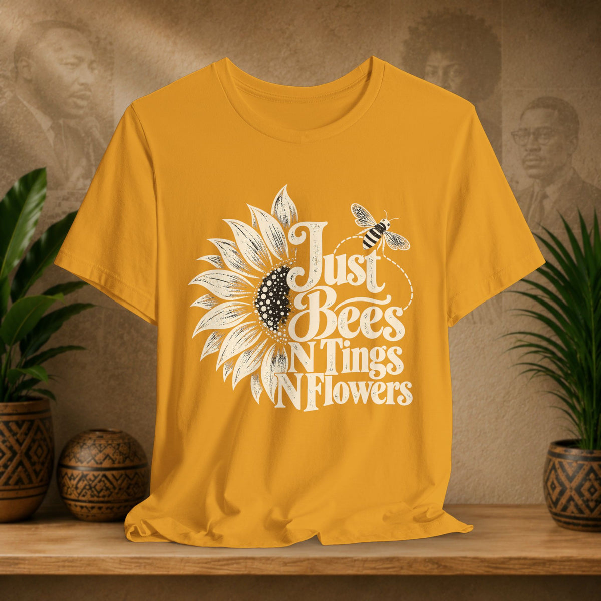 A mustard yellow short-sleeve t-shirt with a white graphic print of a sunflower, a bee, and the text 'Just Bees N Tings N Flowers', displayed on a wooden shelf with potted plants and a textured background.