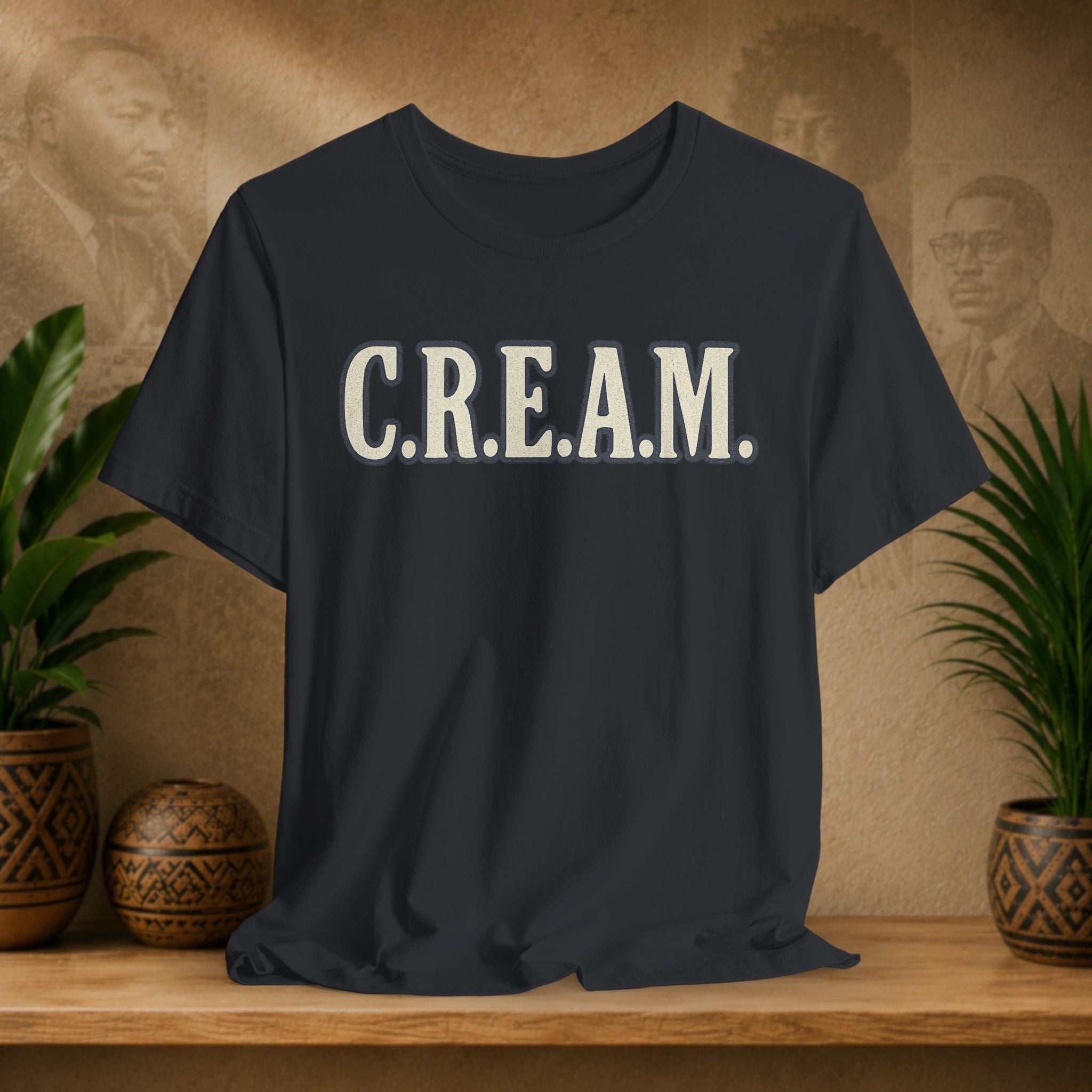 Black t-shirt with 'C.R.E.A.M.' text on a wooden surface with plants and a textured wall background