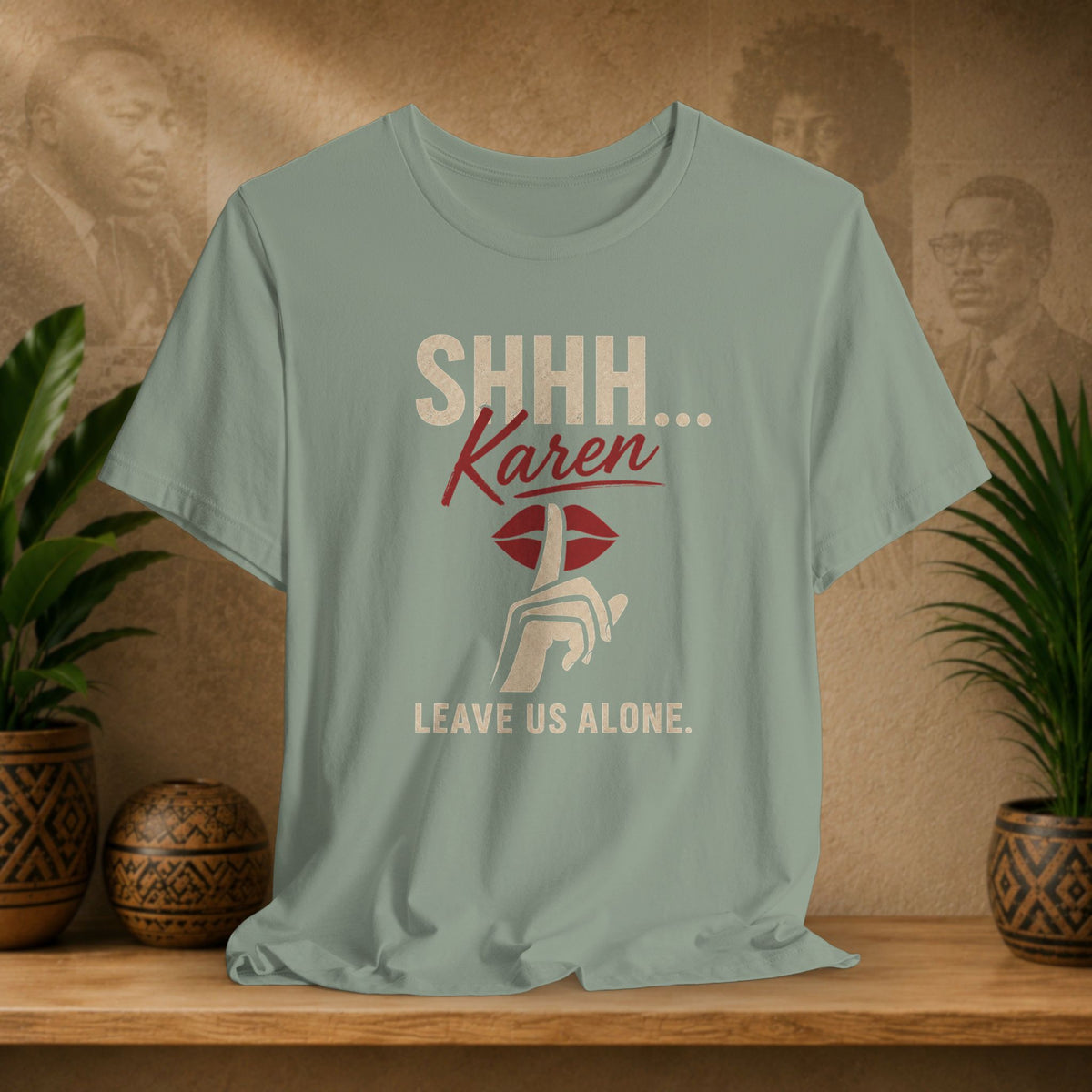 A sage green graphic t-shirt displayed on a wooden shelf with potted plants and a textured background. The shirt features a graphic with the text 'SHHH.