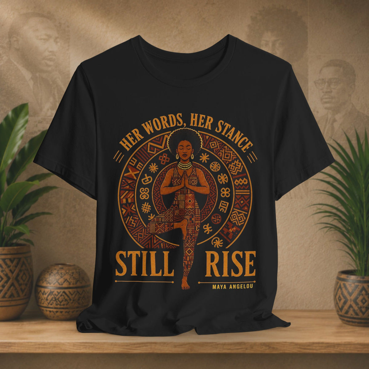 A black short-sleeve graphic T-shirt displayed on a wooden surface against a warm beige wall with decorative plants. The shirt features a large front print with an illustrated figure in a meditative pose inside a circular design, with the text 'HER WORDS, HER STANCE STILL RISE' and 'MAYA ANGELOU' below.