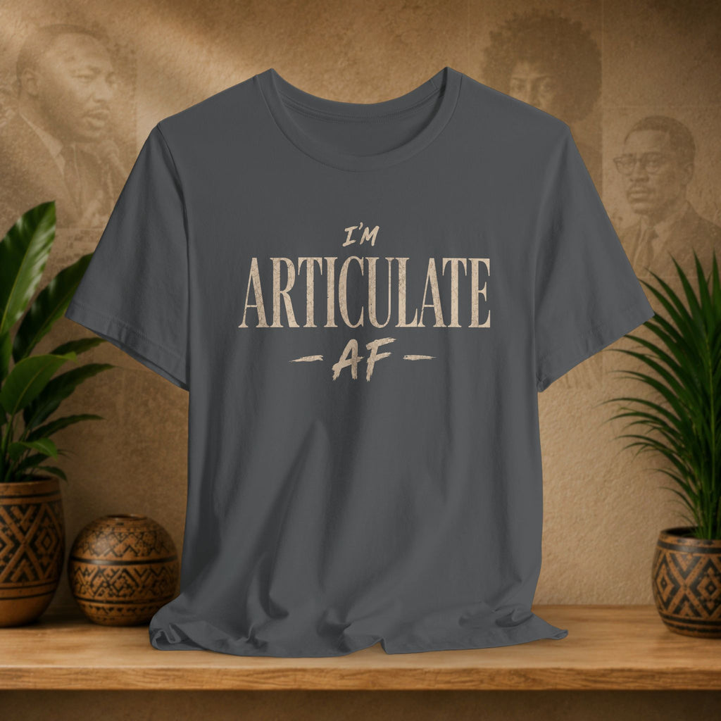 A dark gray crewneck t-shirt with the text 'I'M ARTICULATE AF' printed in a distressed beige font, displayed on a wooden shelf with potted plants and a textured background.