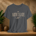 A dark gray crewneck t-shirt with the text 'I'M ARTICULATE AF' printed in a distressed beige font, displayed on a wooden shelf with potted plants and a textured background.