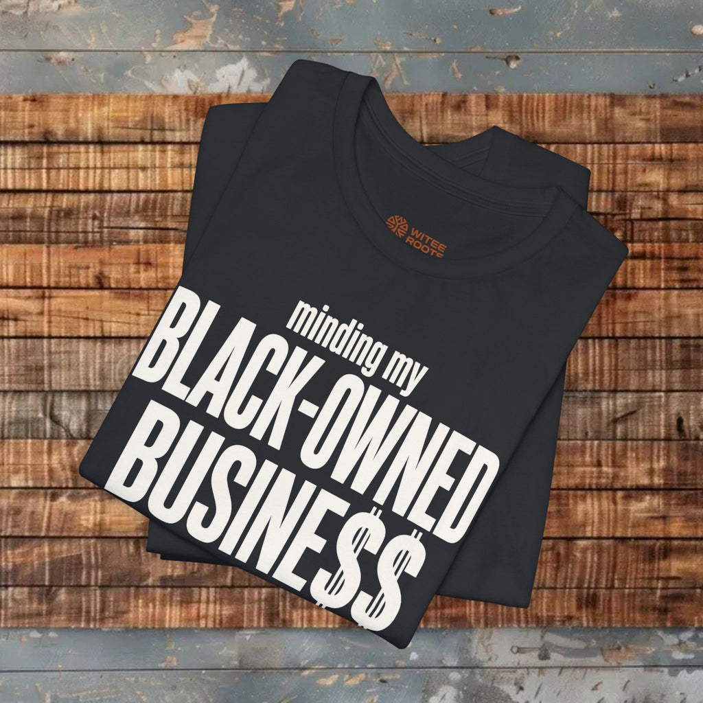 A folded black t-shirt with bold white text reading 'minding my BLACK-OWNED BUSINESS$' is displayed on a rustic wooden plank surface.