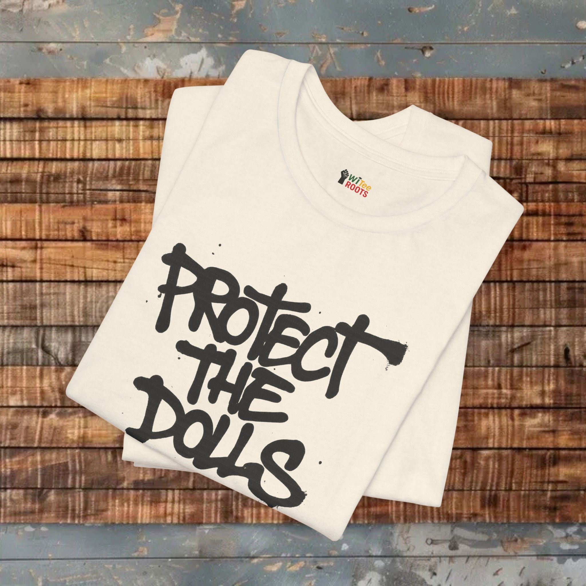 White t-shirt with 'Protect the Dolls' text on a wooden surface