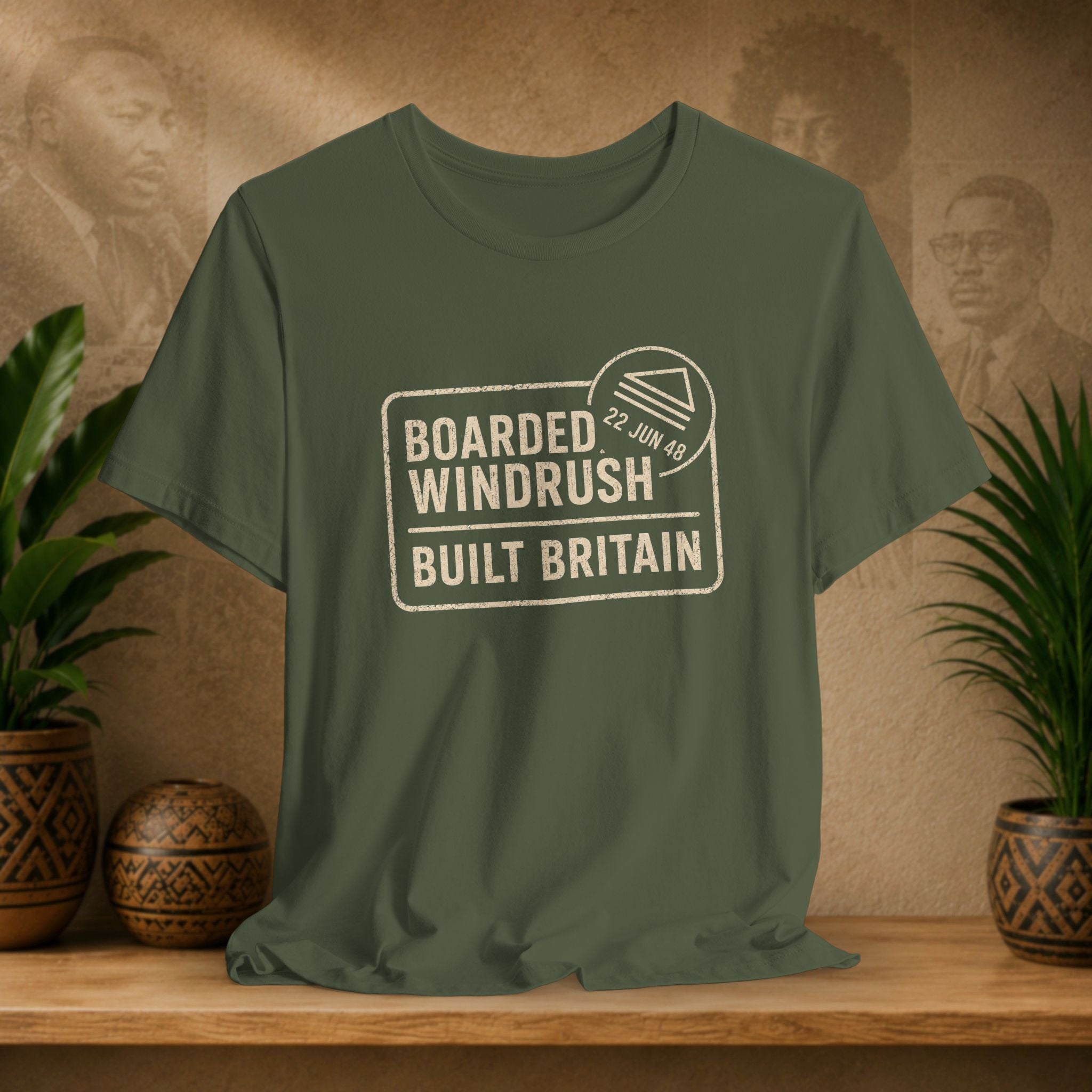 Green t-shirt with 'Boarded Windrush Built Britain' text on a wooden surface with plants and a textured wall background.