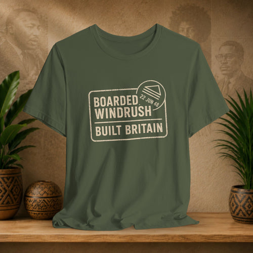 Green t-shirt with 'Boarded Windrush Built Britain' text on a wooden surface with plants and a textured wall background.
