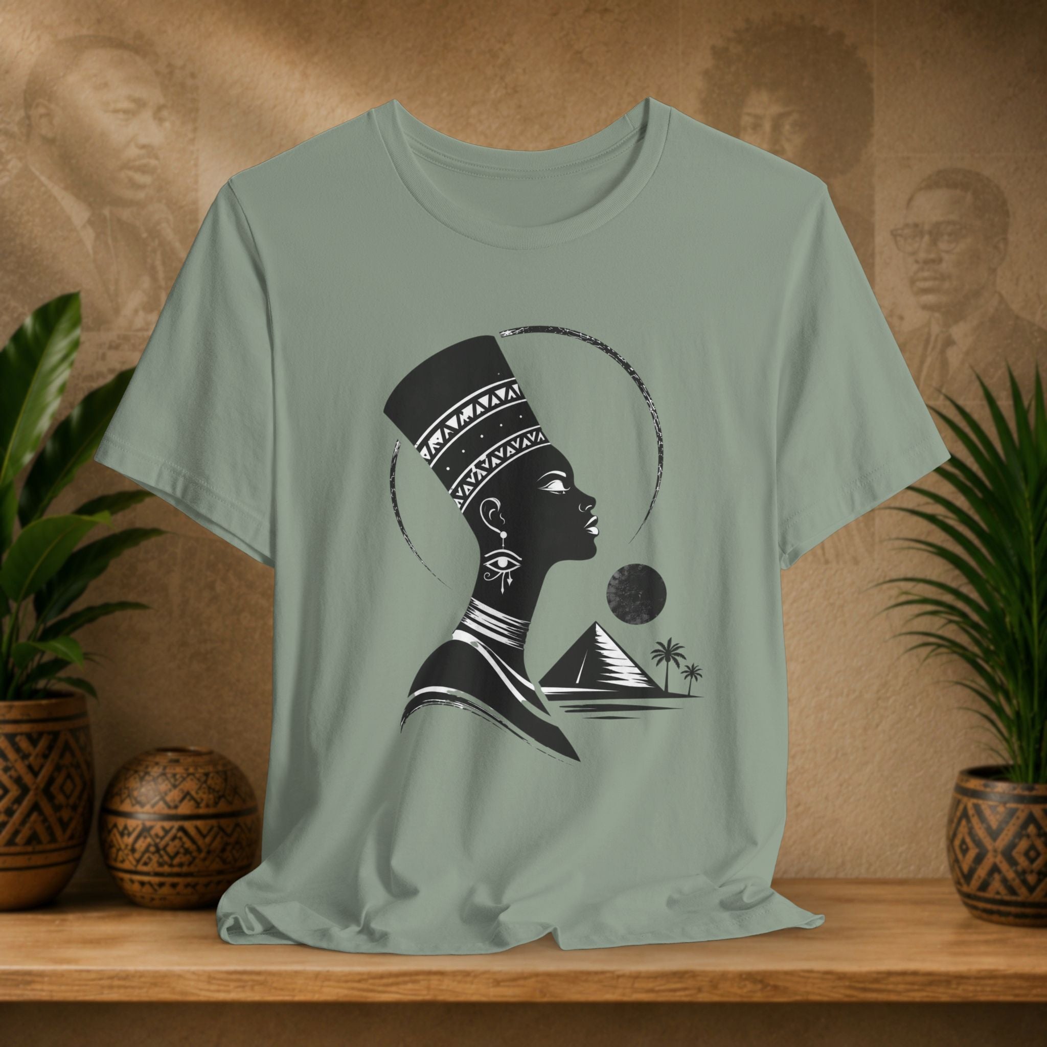 Sage green t-shirt with a black silhouette design of a woman, pyramid, and palm trees on a wooden surface with plants and decorative items.