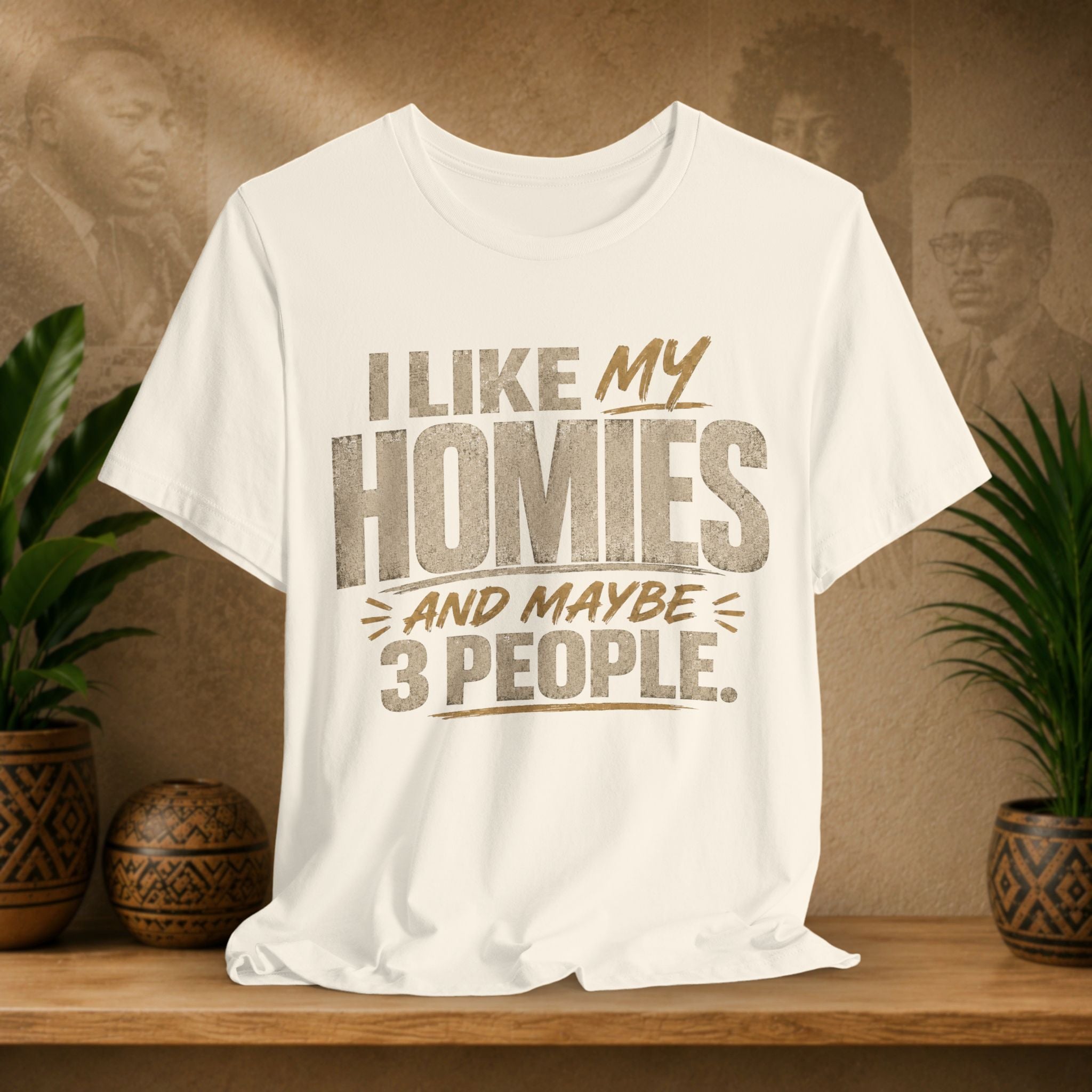A beige graphic t-shirt displayed on a wooden shelf. The shirt features a humorous slogan printed in white and tan lettering: 'I LIKE MY HOMIES and maybe 3 PEOPLE.